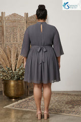 Back view of a plus size woman wearing a grey pleated high neck short dress with a keyhole back opening and a tied waist belt.