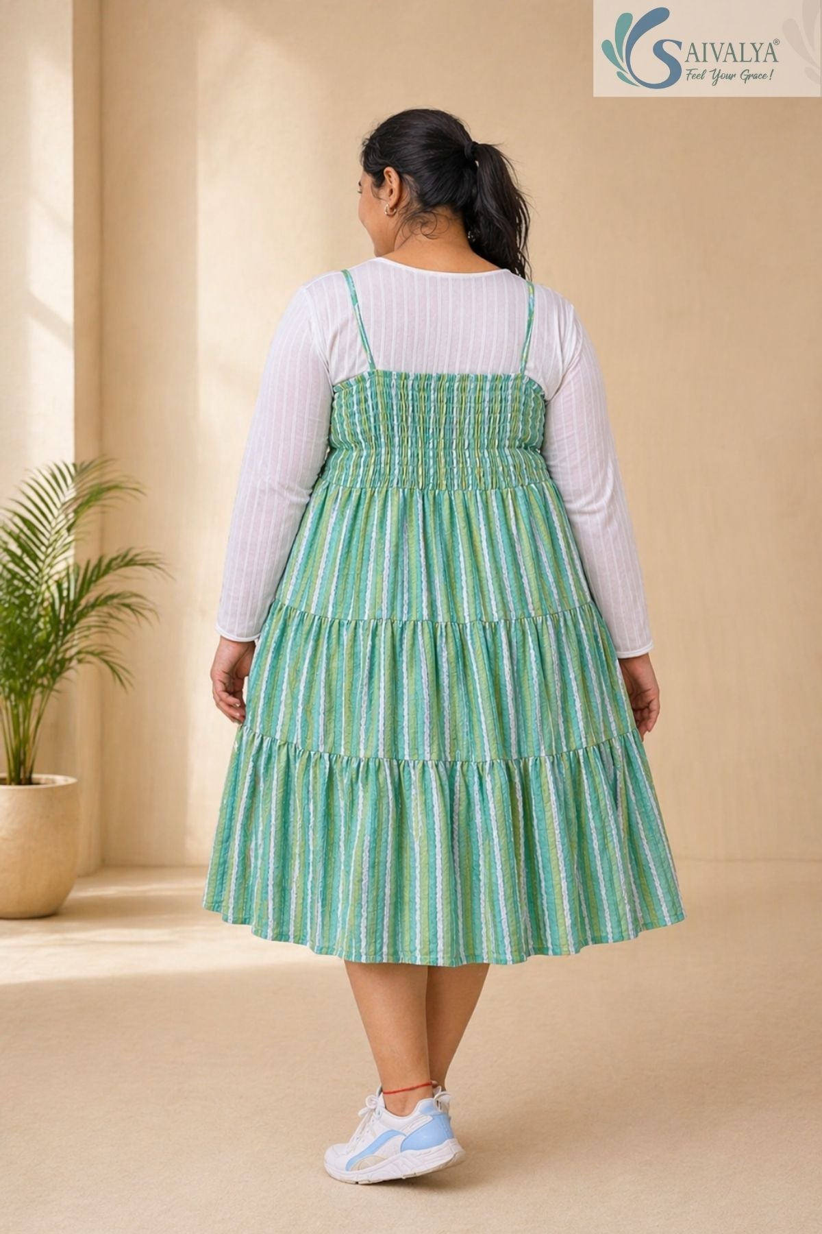 Back view of a woman wearing the Saivalya Verdant Muse green striped tiered midi dress layered over a white long-sleeve top showcasing the smocked shirred back detail paired with sneakers