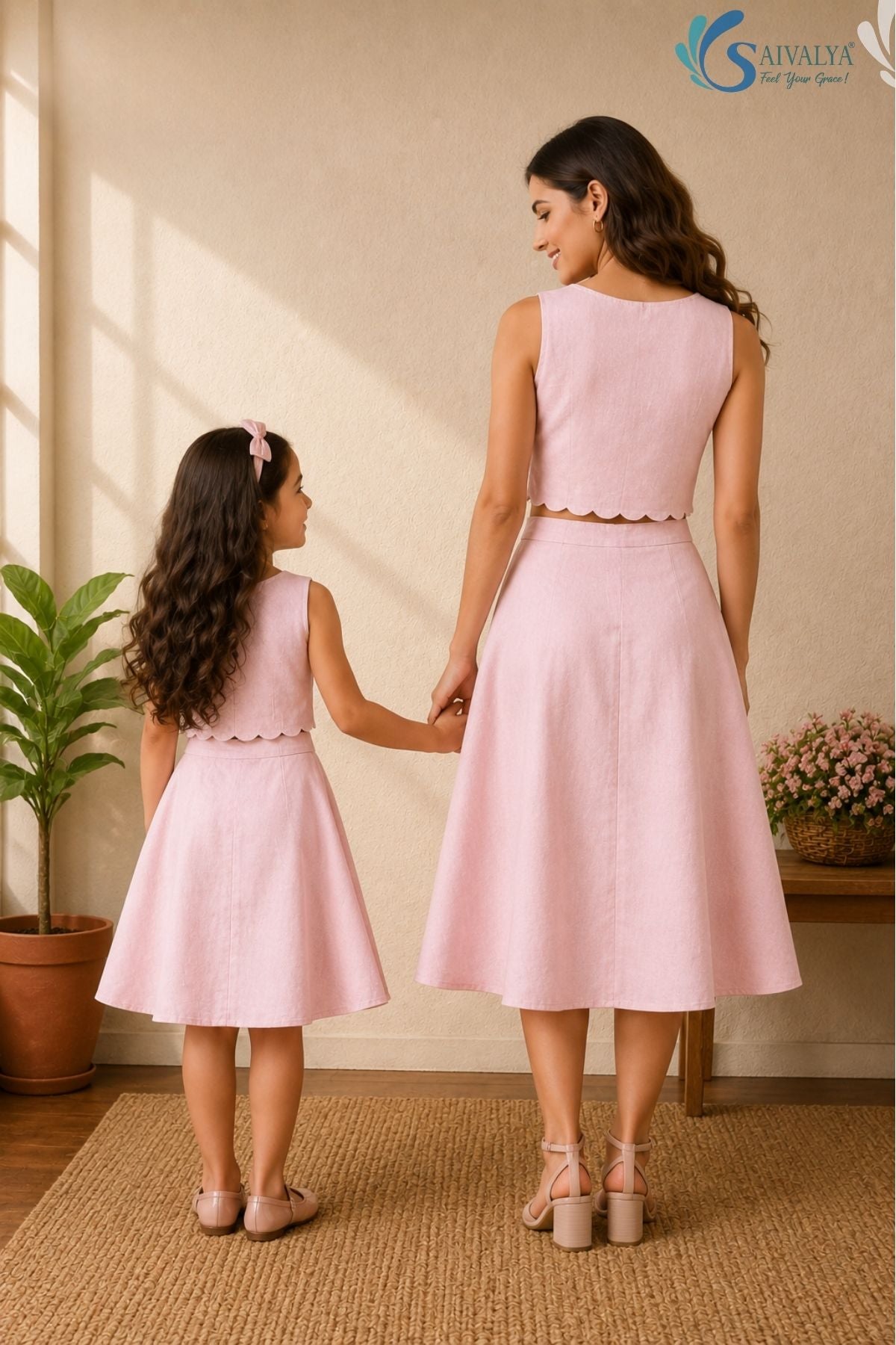 Full-length back view of a woman and a young girl holding hands while standing facing away from the camera in a bright, warm indoor studio setting with a woven rug, a potted fiddle-leaf fig plant, and a wooden bench in the background; both are wearing matching powder pink cotton co-ord sets showcasing the smooth back panels of the sleeveless crop tops with scalloped hems and the structured lines of the high-waisted, flared A-line skirts.