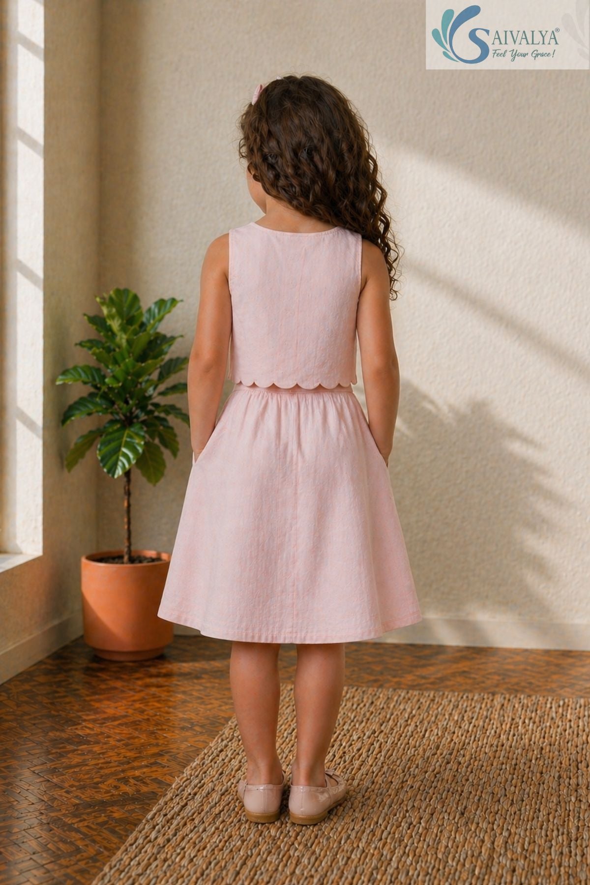 Back view of a young girl standing with her hands in her pockets, wearing the Blush Petal Cotton Co-ord set featuring a sleeveless crop top with a scalloped hem and a matching elastic-back A-line skirt.