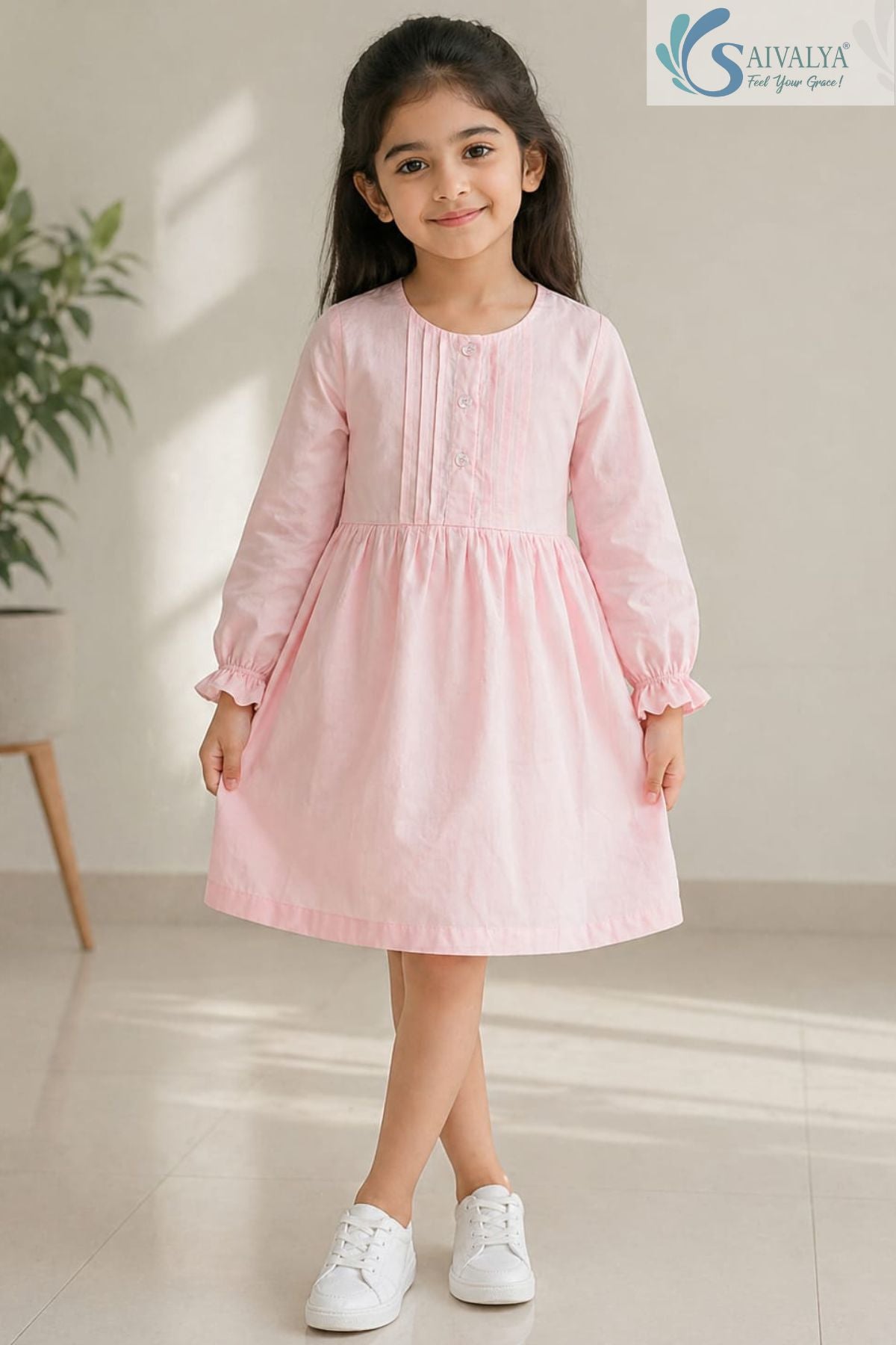 Front view of a smiling young girl with legs crossed wearing the Blush Pink Pin-Tuck Cotton Frock featuring a gathered waist, long sleeves with ruffled cuffs, vertical pin-tuck detailing on the bodice, and white sneakers.
