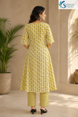 Back view of a woman standing in a sunny, indoor-outdoor setting, wearing the Citrus Blossom Cotton Kalidar Kurta Set showcasing the detailed olive-yellow floral print across the flared anarkali kurta, paired with geometric print straight pants and black low heels.
