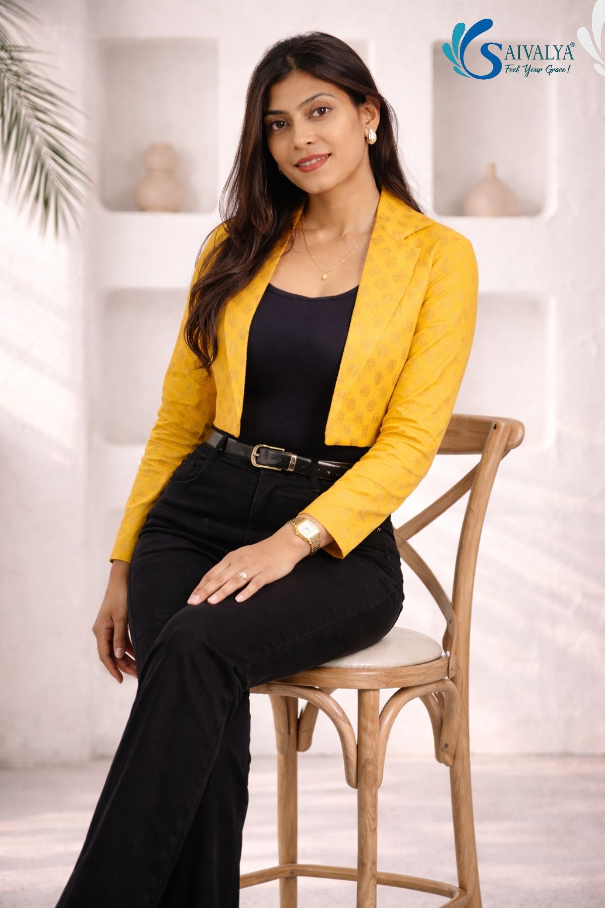 Three-quarter full-length view of a woman smiling while sitting on a wooden high chair in a bright studio setting with wall niches and a plant in the background, wearing the Gold Jacquard Cropped Blazer featuring its structured short silhouette and self-patterned texture, layered over a black scoop-neck top and paired with high-waisted black trousers with a black leather belt.