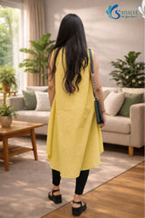 Full-length back view of a woman standing in a cozy living room setting with a sofa and plants, wearing the Ira Mustard High-Low Kurta highlighting the back profile and asymmetric high-low hemline of the yellow sleeveless kurta, paired with black leggings and black platform sandals.