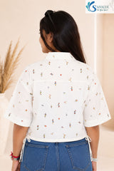 Medium back view of a woman standing in a bright studio setting with pampas grass in the background, wearing the Ivory Micro-Print Drawstring Shirt highlighting its structured back yoke, short wide sleeves, traditional collar, scattered colorful miniature graphics, and side drawstring ties at the hem, paired with blue denim jeans.