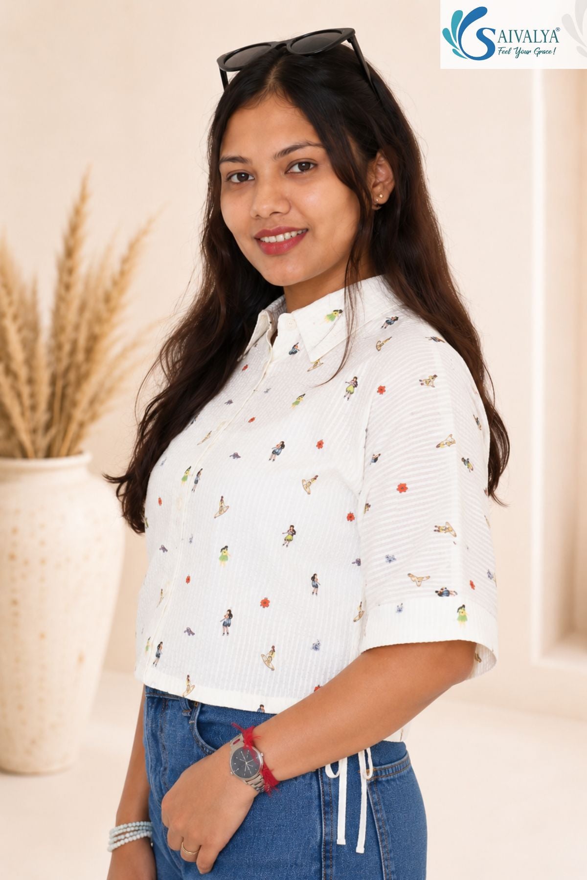 Medium three-quarter side view of a smiling woman posing in a bright studio setting with a large decorative vase in the background, wearing the Ivory Micro-Print Drawstring Shirt showcasing its short wide sleeves, traditional collar, scattered colorful miniature graphics, and side drawstring tie details at the hem, paired with blue denim jeans.