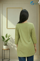 Full-length back view of a woman standing in a minimalist indoor studio setting with a gold frame and a potted plant in the background, wearing the Prakriti Sage Overlap Tunic highlighting its clean back profile, round neckline, and three-quarter sleeves, paired with blue denim jeans.