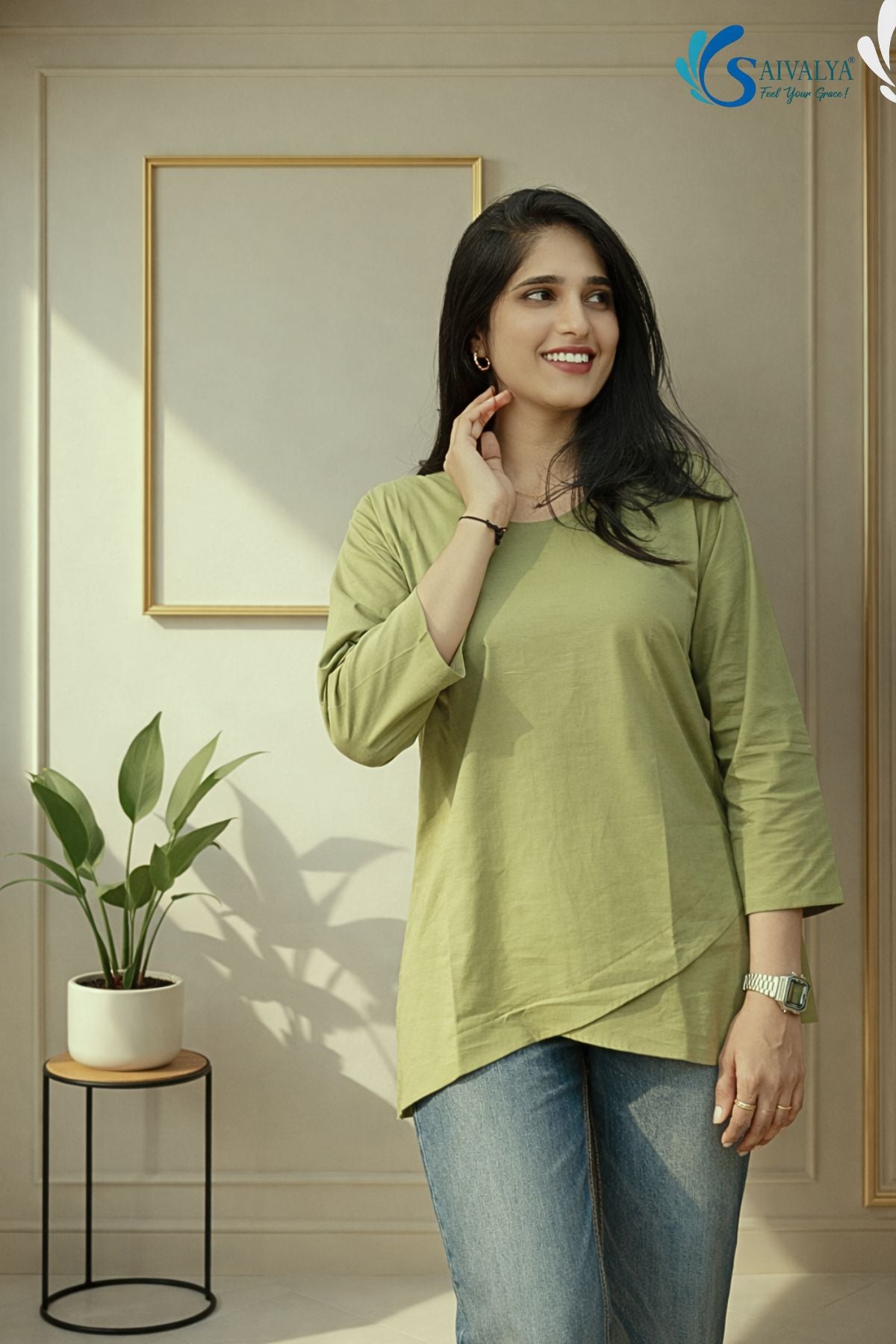 Three-quarter medium view of a smiling woman looking off-camera with a hand resting near her neck in a minimalist indoor studio setting with a gold frame and a potted plant in the background, wearing the Prakriti Sage Overlap Tunic highlighting its round neckline, three-quarter sleeves, and asymmetric overlap tulip hemline, paired with blue denim jeans.