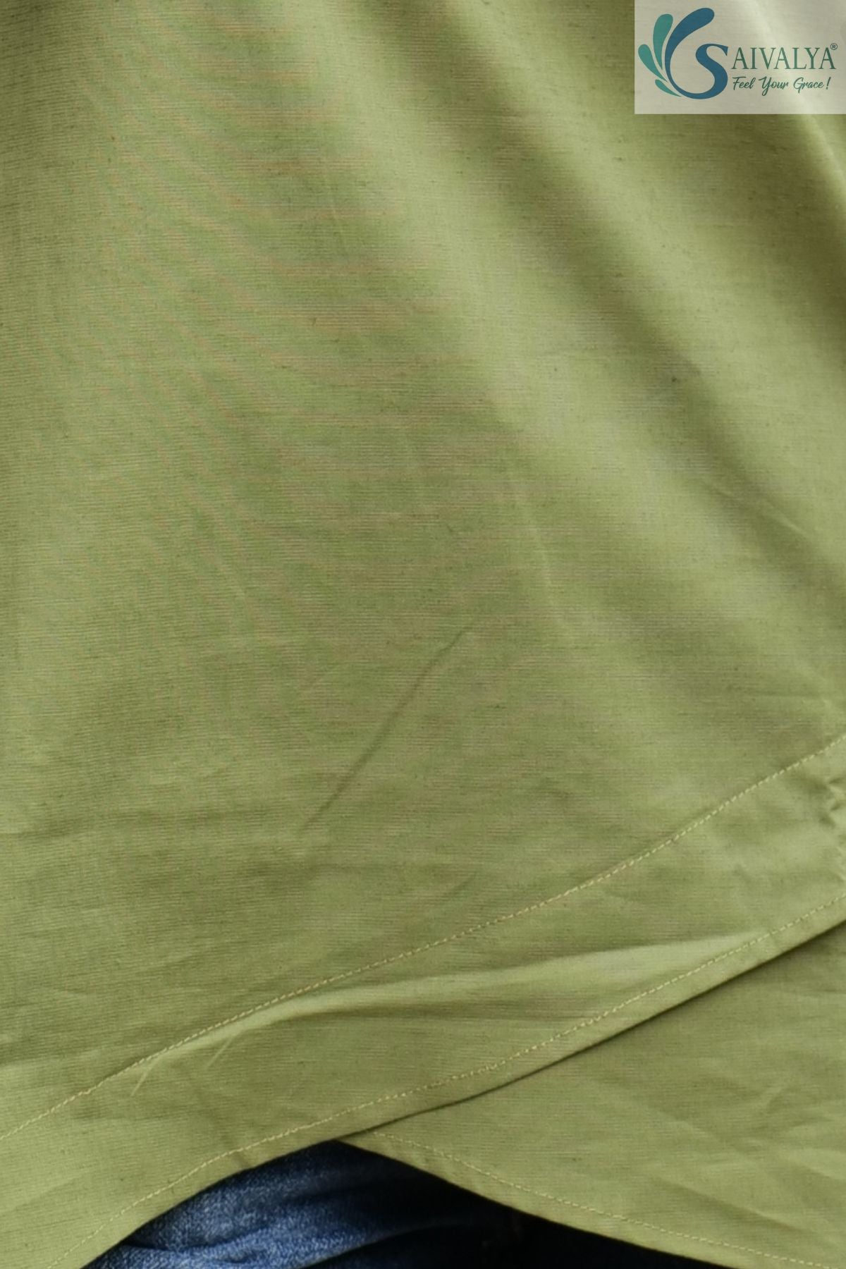 Close-up macro view highlighting the smooth light sage green cotton-blend fabric from the Prakriti Sage Overlap Tunic, showcasing the fine texture and the curved stitched panels of the asymmetric overlap tulip hemline.