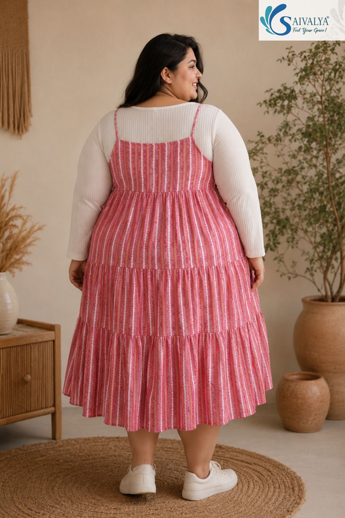 Back view of a woman standing on a round jute rug in a cozy bohemian room, looking over her shoulder, wearing the Raspberry Striped Tiered Midi Dress layered over a ribbed off-white long-sleeve top, highlighting the straight back neckline, thin straps, and pink striped tiered flared skirt, paired with white sneakers.