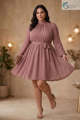Full-length front view of a woman standing gracefully with legs crossed and holding the hem of her skirt, wearing the rosewood Pleated Muse Dress featuring a high neck, crinkle pleating, long balloon sleeves, and a self-tie belt.