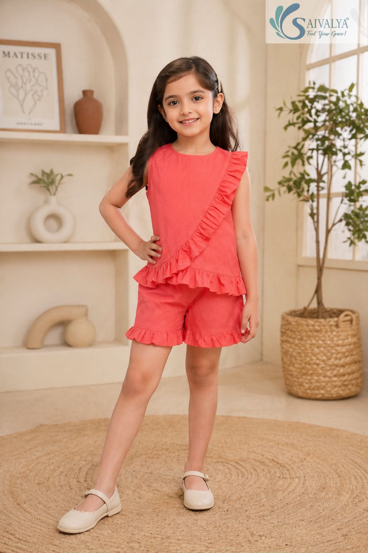 Full-length front view of a smiling young girl posing with her hand on her hip, wearing the coral pink Ruffle Charm Co-ord Set featuring a sleeveless asymmetric top with diagonal ruffle detailing and matching ruffle-trim casual shorts.