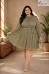 Full-length front view of a smiling plus-size woman posing gracefully in a warm, elegantly styled indoor setting with a wooden console table and potted palm tree in the background, wearing the Sage Georgette Pleated Muse Dress featuring a light olive-sage green plissé pleated fabric, a high mock neckline, long sheer sleeves with ruffled cuffs, a matching fabric waist tie, and a flared pleated skirt, paired with nude pointed heels and a beige clutch.