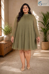 Full-length front view of a smiling plus-size woman standing facing forward in a warm, elegantly styled indoor setting with a wooden console table and potted palm tree in the background, wearing the Sage Georgette Pleated Muse Dress styled without its belt to showcase a loose, flowing babydoll silhouette in light olive-sage green plissé pleated fabric with a high mock neckline and long sheer sleeves with ruffled cuffs, paired with nude pointed heels and a beige clutch.
