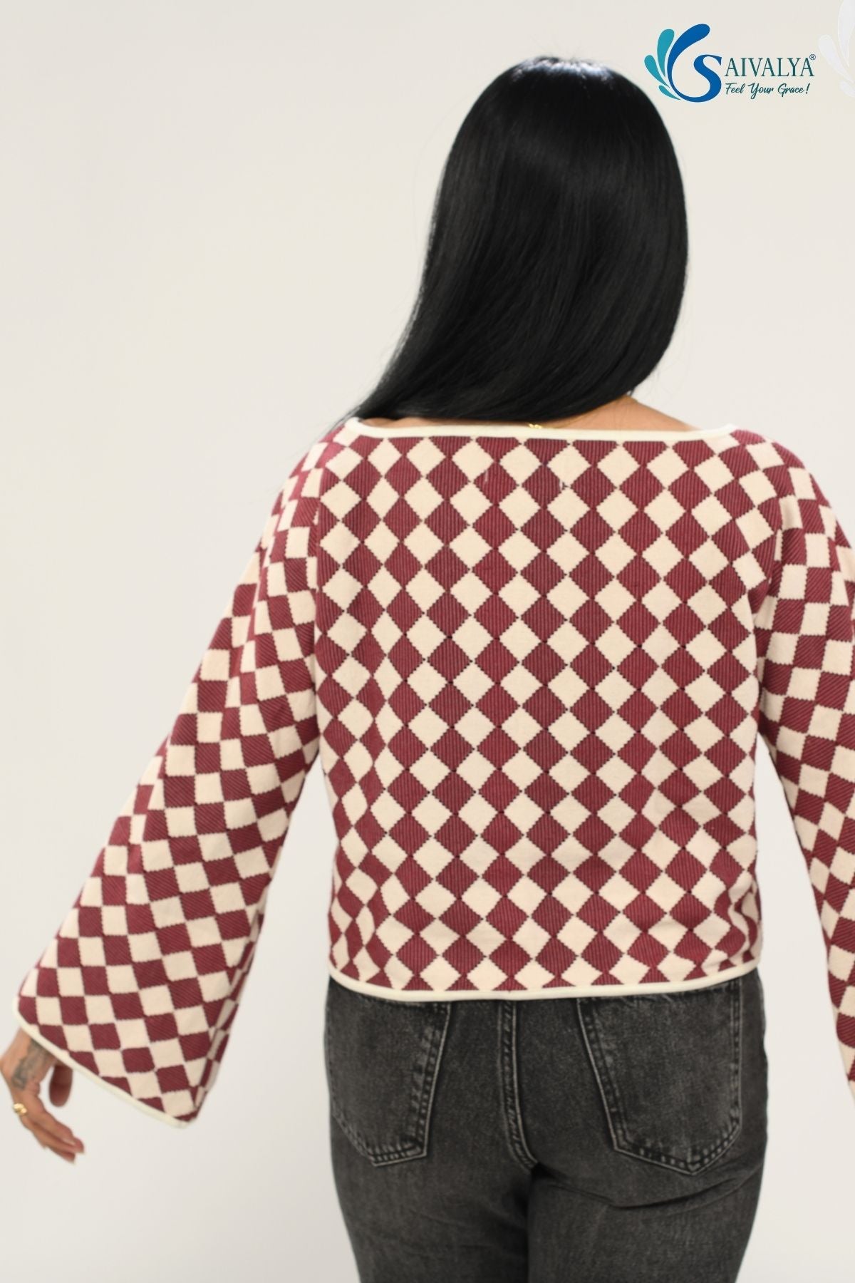 Rear view of the Shatranj Maroon Bell Top, showcasing the continuous maroon and cream harlequin checkered pattern across the back and wide bell sleeves. The top features a clean boat neckline with cream piping, paired with dark charcoal grey jeans.