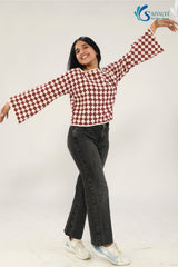 Full-length view of a woman smiling and posing with arms outstretched to show the dramatic flare of the Shatranj Maroon Bell Top. She is wearing the maroon and cream checkered top with high-waisted dark grey wide-leg jeans and white sneakers against a bright studio background.