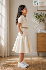 Side view of a smiling young girl standing with her hand in her pocket, wearing a cream Slub Cotton Co-ord set featuring a short-sleeve collared crop shirt and a matching flared midi skirt.