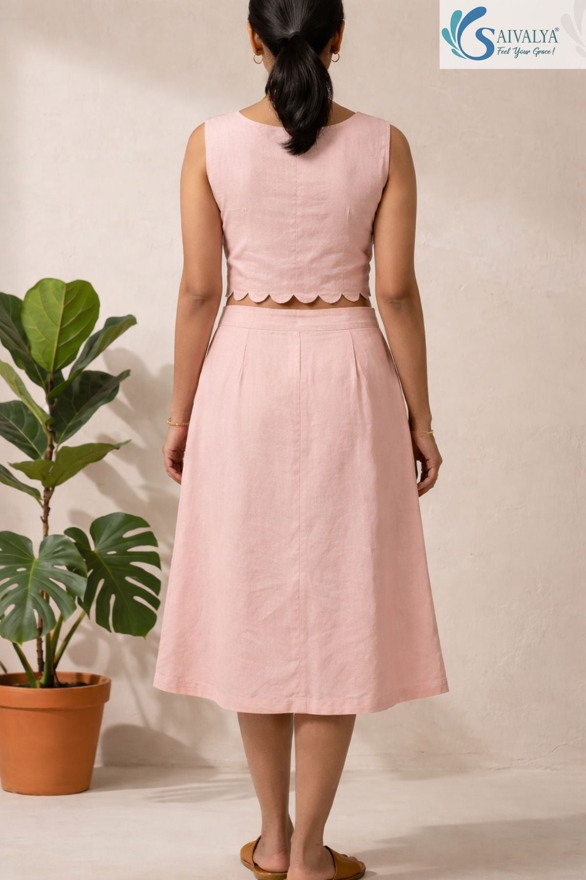 Full-length back view of a woman standing facing away from the camera in a bright, minimalist indoor studio setting with a potted fiddle-leaf fig plant in the background, wearing The Scalloped Blush Midi Set highlighting its sleeveless powder pink cotton crop top with a scalloped petal-cut hemline, a central vertical back seam, and a matching high-waisted, A-line midi skirt with subtle back pleating details, completed with tan leather flat slides.