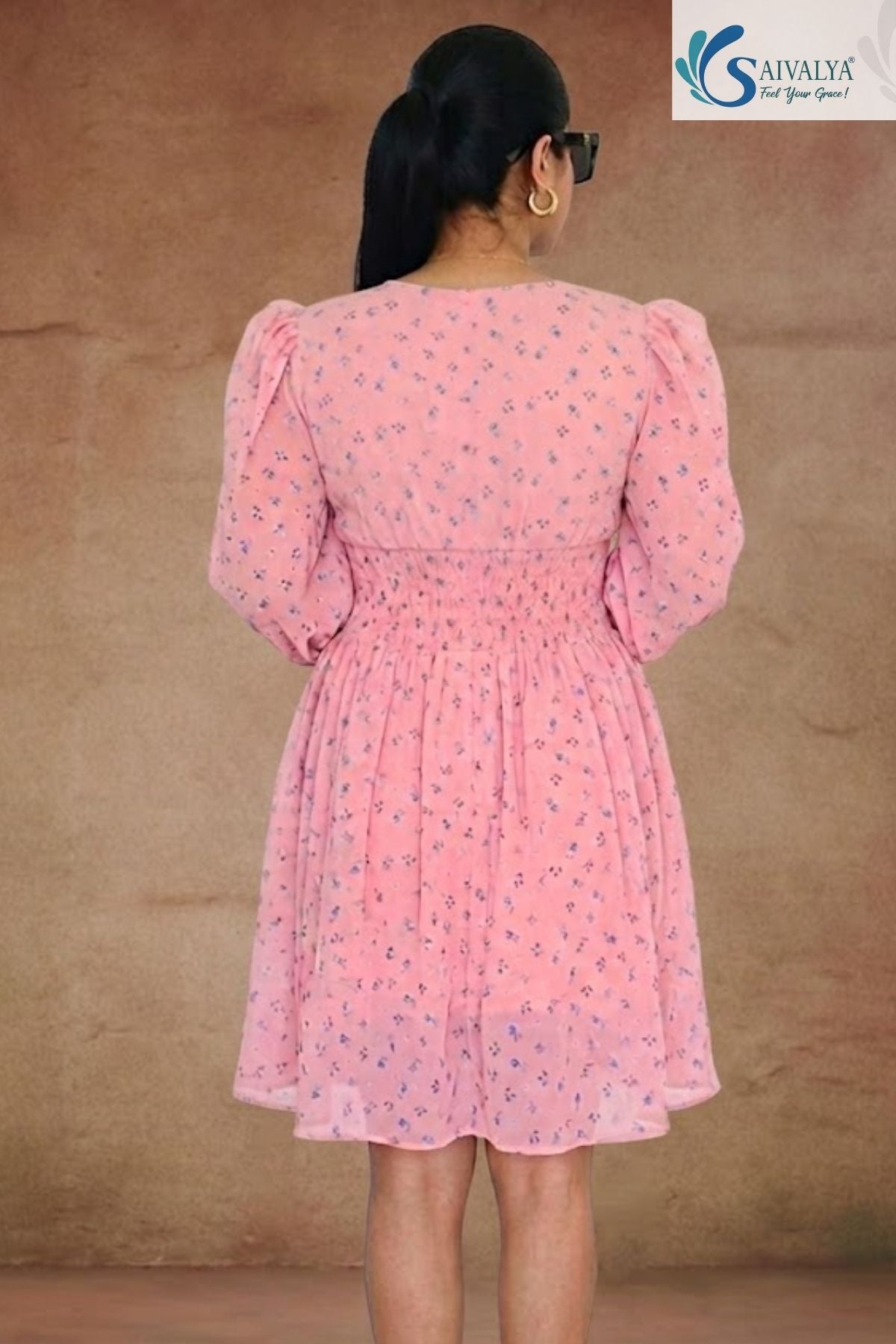 Full-length back view of a woman standing facing away from the camera in a warm, textured indoor studio setting, wearing the Aashni Smocked Fit & Flare Dress highlighting its light pink georgette fabric with blue micro-floral prints, structured back yoke, gathered puff shoulders, and a wide smocked elastic waistband.