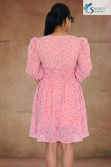 Full-length back view of a woman standing facing away from the camera in a warm, textured indoor studio setting, wearing the Aashni Smocked Fit & Flare Dress highlighting its light pink georgette fabric with blue micro-floral prints, structured back yoke, gathered puff shoulders, and a wide smocked elastic waistband.