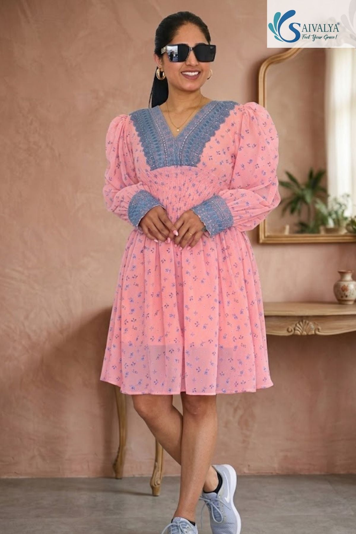 Full-length front view of a smiling woman posing with crossed legs in a warm, textured indoor studio setting with a wooden console table and mirror in the background, wearing the Aashni Smocked Fit & Flare Dress featuring a light pink georgette fabric with blue micro-floral prints, a V-neckline adorned with thick dusty blue embroidered borders, long puff sleeves with matching embroidered cuffs, and a smocked elastic waistband, paired with black sunglasses and grey sneakers.