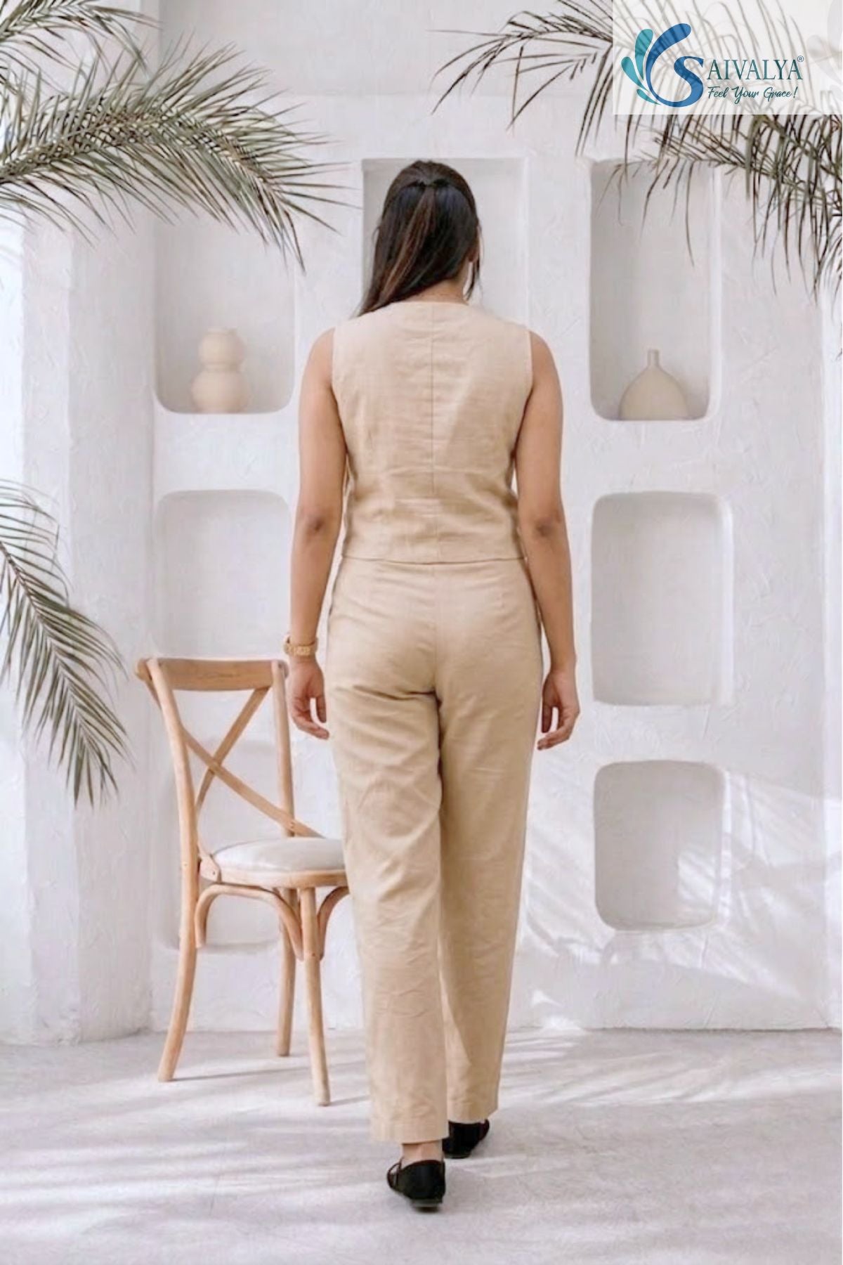 Rear view of a woman wearing the beige Aatman Waistcoat and Straight Trousers Set. The image showcases the sleeveless back of the waistcoat and the clean, tailored fit of the matching straight-leg trousers, set against a minimalist white studio background with decorative wall alcoves.