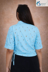Rear view of a woman wearing the Azure Sky Printed Drawstring Shirt, showcasing the continuous light blue seersucker pattern and miniature illustrative prints across the back. The image highlights the cuffed short sleeves and the adjustable drawstring tie visible on the left side hem.