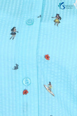 Alt Text:A macro close-up of the Azure Sky Printed Drawstring Shirt, highlighting the light blue seersucker fabric with its distinct puckered, checkered texture. The image shows matching blue buttons and detailed miniature prints of surfers, hula dancers, and tropical flowers.
