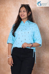A woman smiling and posing in the Azure Sky Printed Drawstring Shirt, a light blue button-down with a subtle checkered texture and small floral prints. The shirt features elbow-length sleeves and adjustable drawstring ties at the hem, styled with black trousers against a textured beige background.