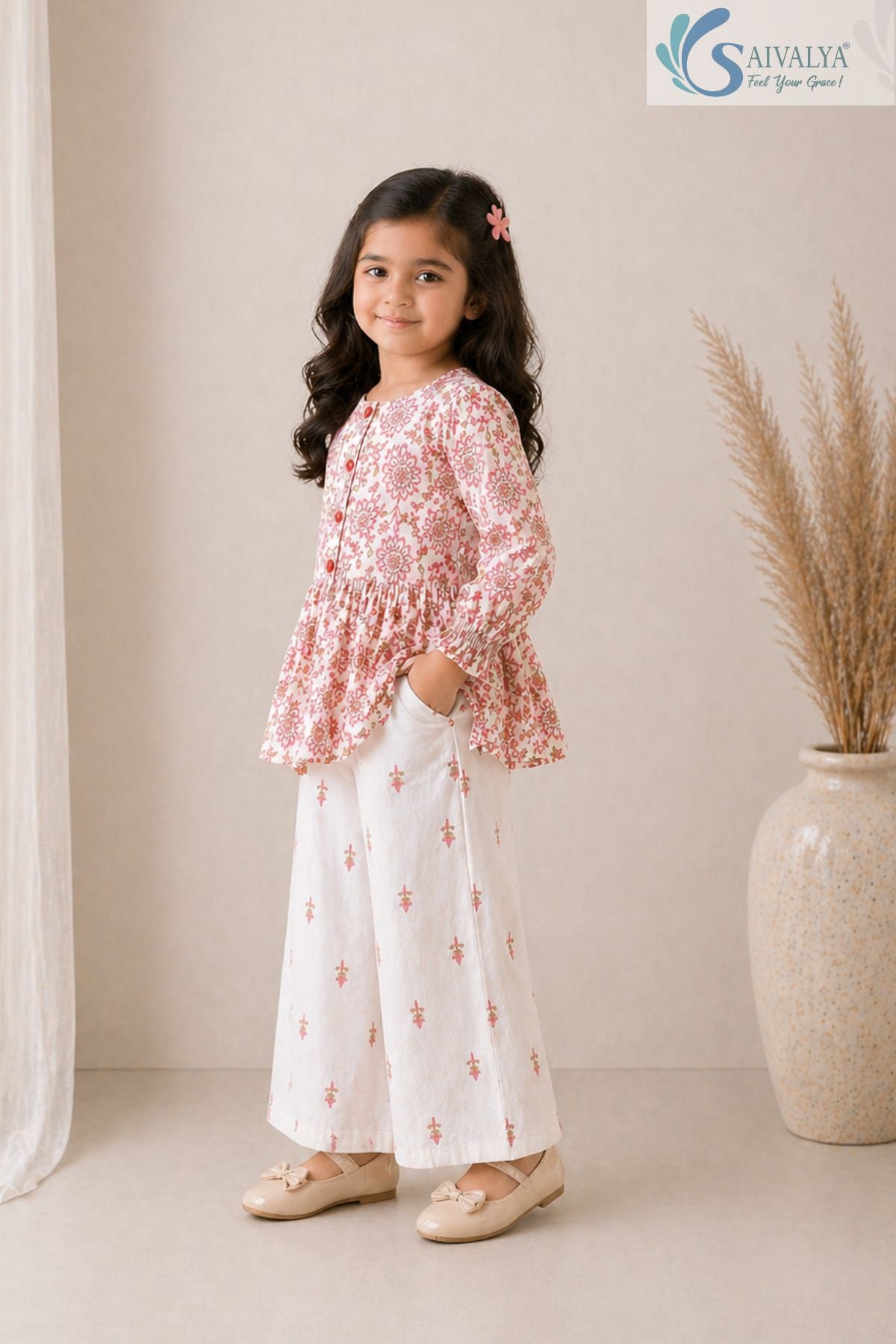 Side view of a young girl standing with her hand in her pocket, wearing the Bloom and Breezy cotton co-ord set with a pink patterned peplum top and printed white trousers.