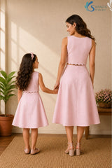 Full-length back view of a woman and a young girl holding hands while standing facing away from the camera in a bright, warm indoor studio setting with a woven rug, a potted fiddle-leaf fig plant, and a wooden bench in the background; both are wearing matching powder pink cotton co-ord sets showcasing the smooth back panels of the sleeveless crop tops with scalloped hems and the structured lines of the high-waisted, flared A-line skirts.