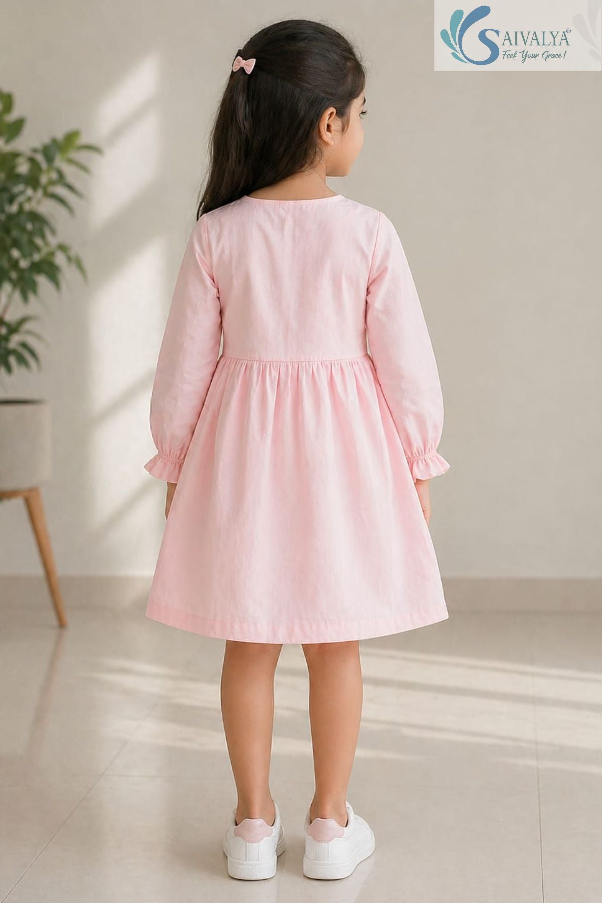 Back view of a young girl standing in a brightly lit room, wearing the Blush Pink Pin-Tuck Cotton Frock featuring a gathered waist, long sleeves with ruffled cuffs, and white sneakers with pink accents.