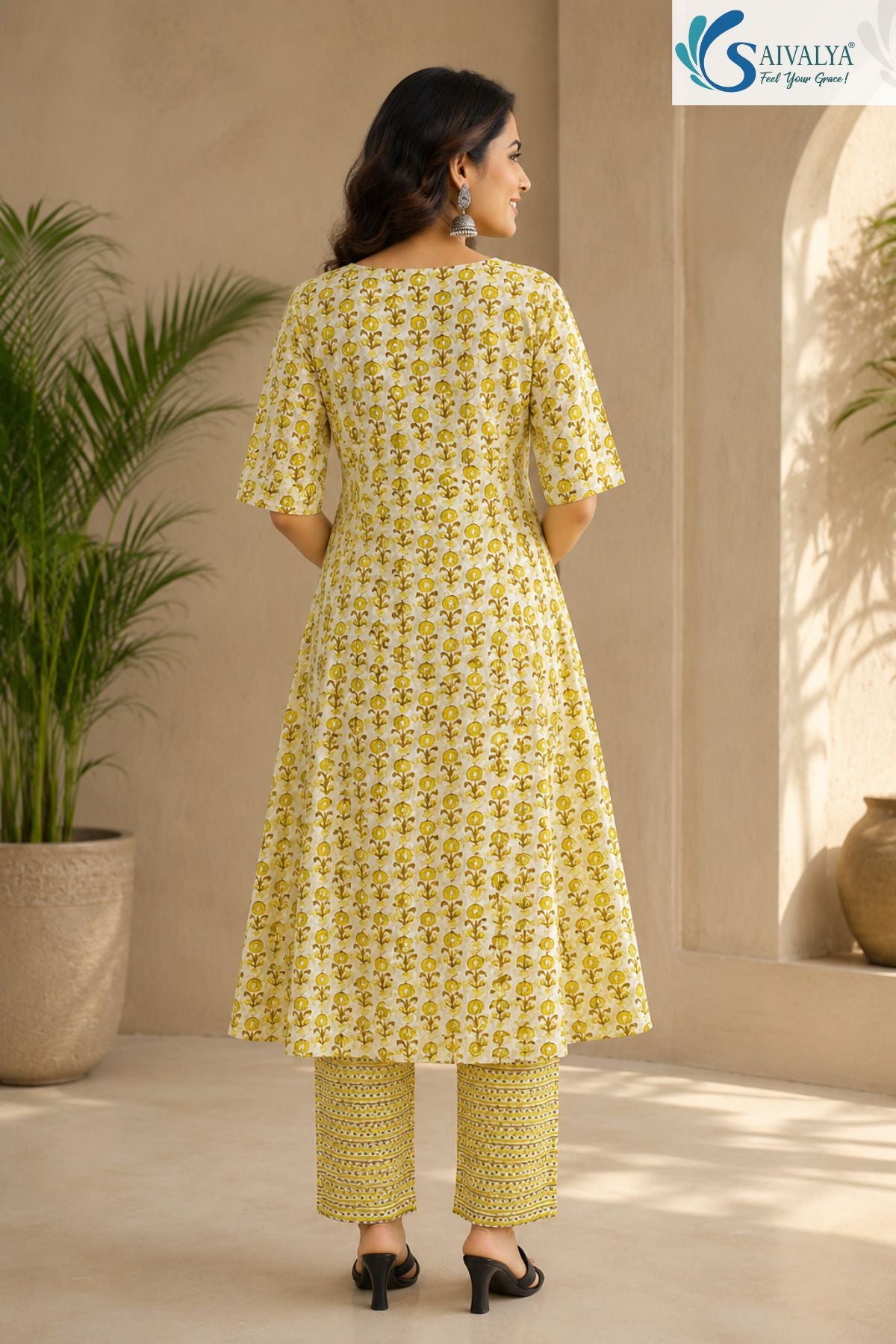 Back view of a woman standing in a sunny, indoor-outdoor setting, wearing the Citrus Blossom Cotton Kalidar Kurta Set showcasing the detailed olive-yellow floral print across the flared anarkali kurta, paired with geometric print straight pants and black low heels.