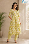 Front view of a smiling woman posing with her hand in her pocket, wearing the Citrus Blossom Cotton Kalidar Kurta Set featuring a white and olive-yellow floral print flared anarkali kurta with elbow-length sleeves, paired with matching geometric print straight pants and black low-heeled sandals.