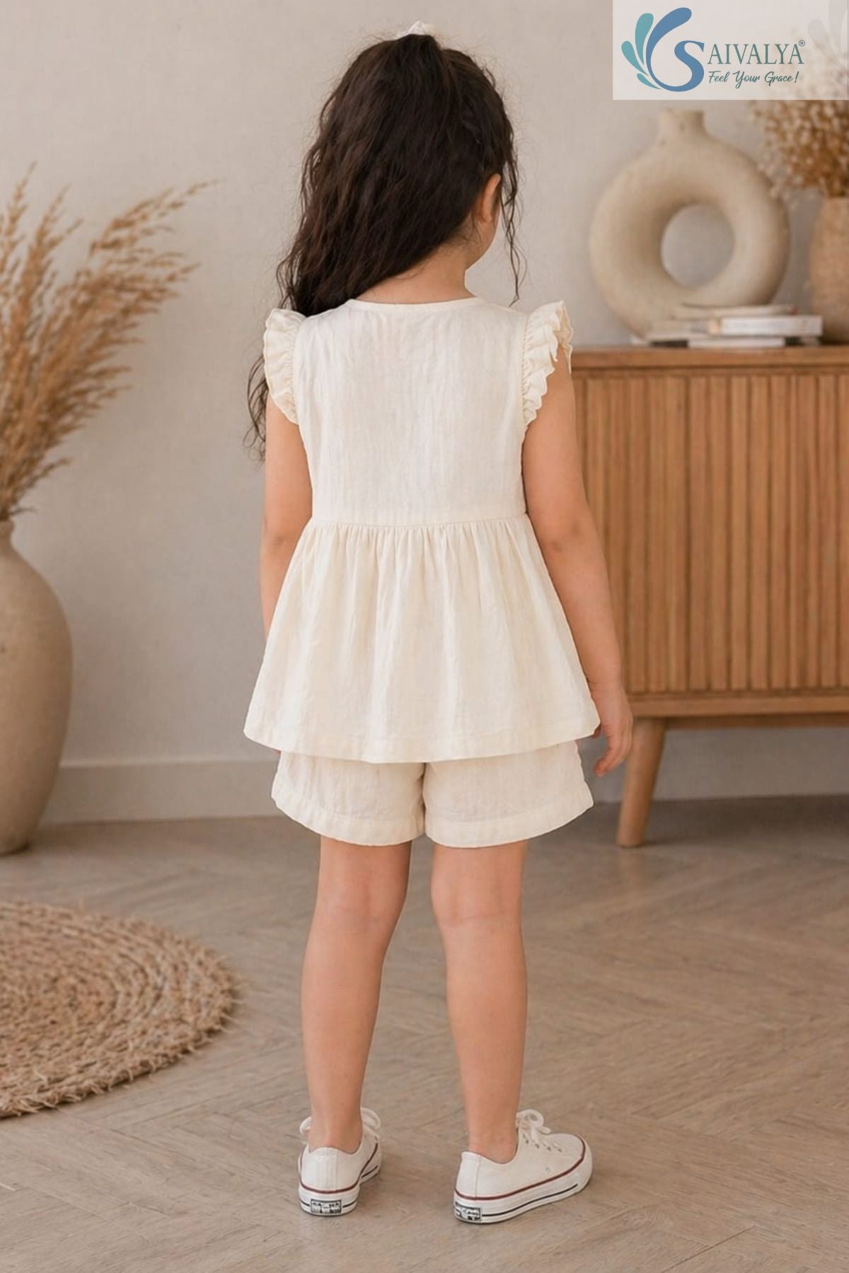 Back view of a young girl standing in a room, wearing the cream Double Gauze Cotton Ruffle Set featuring a sleeveless gathered top with ruffle sleeve details, matching shorts, and white canvas sneakers.