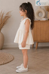 Side view of a smiling young girl standing in a room, wearing the cream Double Gauze Cotton Ruffle Set featuring a sleeveless gathered top with ruffled shoulder straps, matching casual shorts, and white canvas sneakers.
