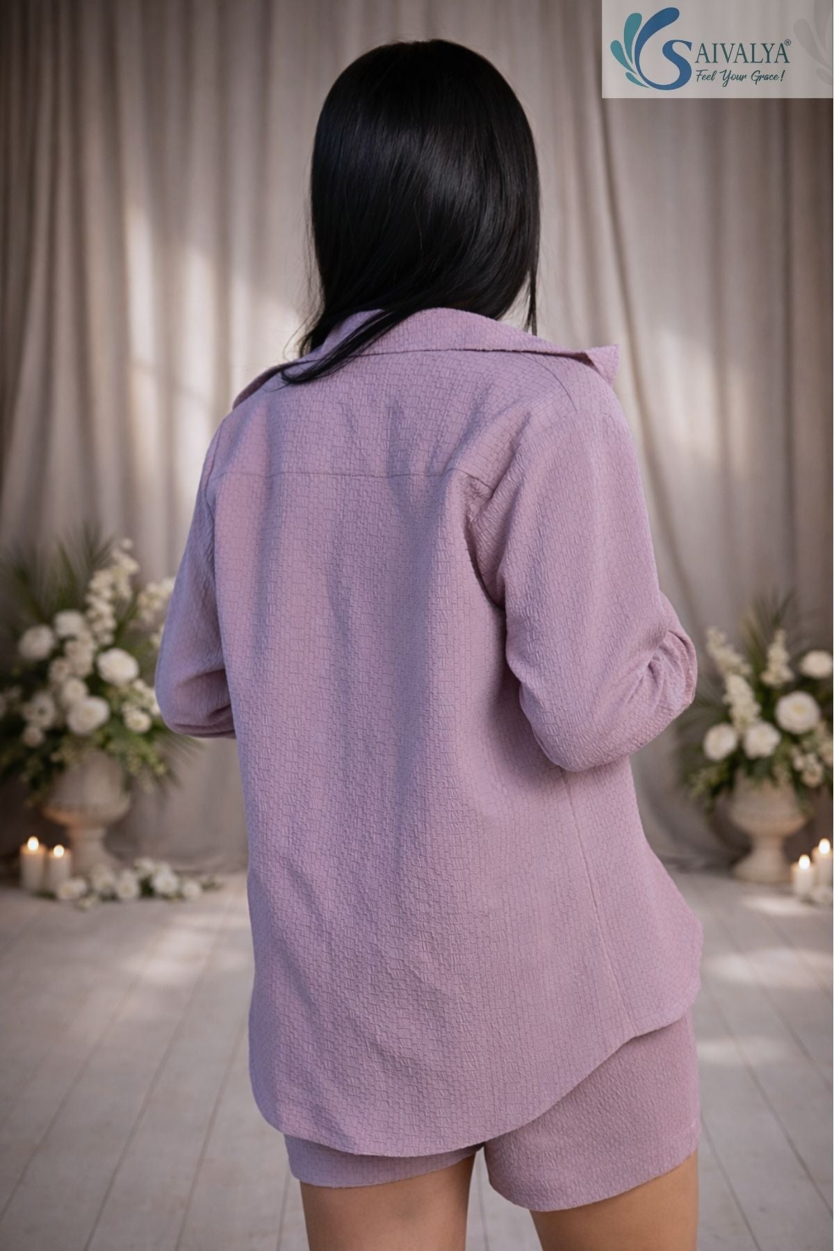 Back view of a young woman standing in a softly lit room with floral arrangements, wearing a Dusty Lavender Textured Shorts Set featuring a long-sleeve collared shirt and matching textured casual shorts.