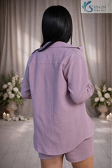Back view of a young woman standing in a softly lit room with floral arrangements, wearing a Dusty Lavender Textured Shorts Set featuring a long-sleeve collared shirt and matching textured casual shorts.