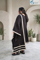 Three-quarter back view of a woman standing in a minimal studio setting with architectural arches and potted plants, wearing the Ebony Monochrome Scalloped 3-Pc Suit highlighting the back profile of the black A-line kurta and a draped black dupatta adorned with bold cream contrast border details.