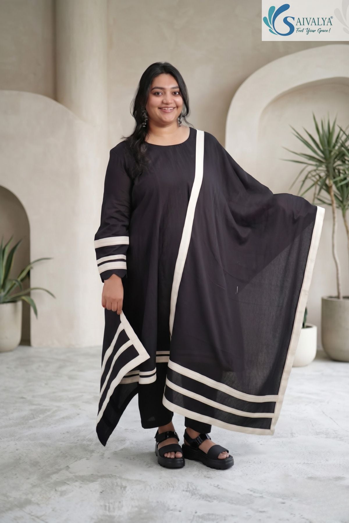 Front view of a smiling woman posing and spreading out her dupatta in a minimal studio setting, wearing the Ebony Monochrome Scalloped 3-Pc Suit featuring a black long-sleeve A-line kurta with cream horizontal border details, matching black pants, and a coordinated black dupatta with wide cream stripes and scalloped sleeve cuffs.