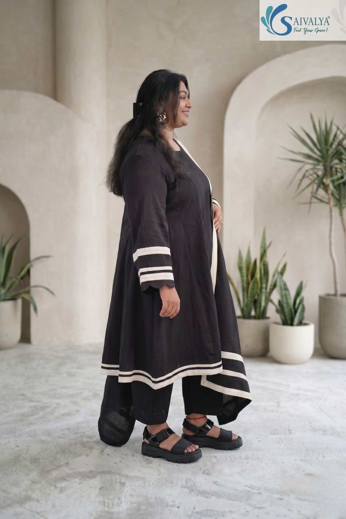 Side view of a smiling woman standing in a minimal studio setting with architectural arches and potted plants, wearing the Ebony Monochrome Scalloped 3-Pc Suit showcasing the side profile of the black flared A-line kurta, matching straight pants, and draped dupatta featuring cream horizontal contrast borders and scalloped sleeve cuffs.
