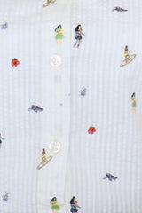 Close-up macro view highlighting the white textured seersucker fabric from the Ivory Micro-Print Drawstring Shirt, showcasing the front button placket with white round buttons and scattered colorful miniature graphics of surfers, hula dancers, and tropical flowers.
