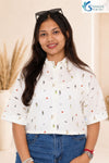 Medium front view of a smiling woman posing in a bright studio setting with pampas grass in the background, wearing the Ivory Micro-Print Drawstring Shirt featuring a white textured button-down design with short wide sleeves, a traditional shirt collar, and colorful miniature graphics scattered throughout, paired with blue denim jeans.