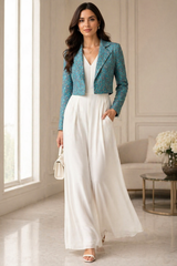 Full-length front view of a woman posing with one hand in her pocket in an elegant, well-lit interior, wearing the Kusum' Short Ethnic Blazer styled over a flowing off-white V-neck wide-leg jumpsuit, highlighting the structured cropped fit and intricate traditional print on a teal base, accessorized with a small white handbag.