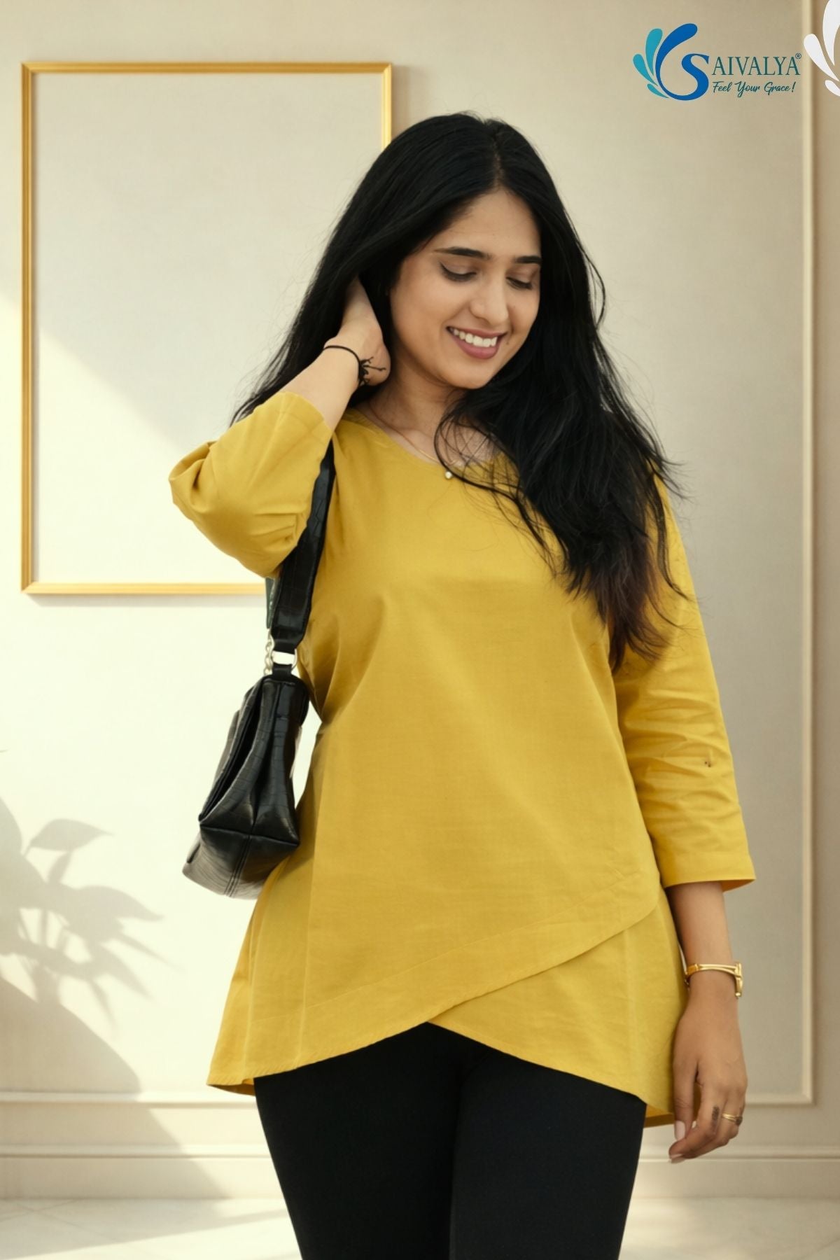 Three-quarter medium view of a smiling woman looking down while running a hand through her hair in a minimalist indoor studio setting with a gold frame in the background, wearing the Ojasvi Mustard Overlap Tunic highlighting its round neckline, three-quarter sleeves, and asymmetric overlap tulip hemline, paired with black leggings and a black shoulder bag.