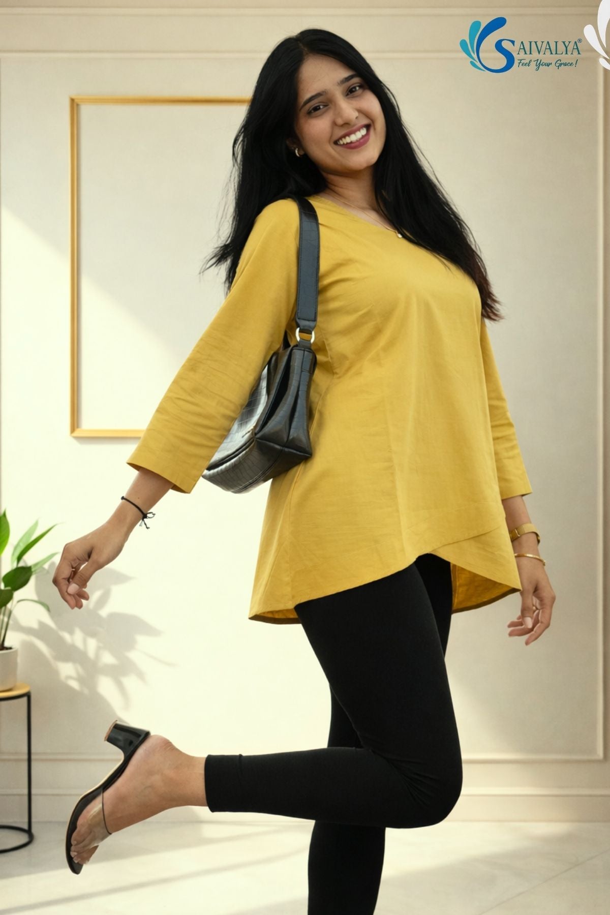 Full-length front view of a smiling woman posing dynamically on one foot in a minimalist indoor studio setting with a gold frame and a potted plant in the background, wearing the Ojasvi Mustard Overlap Tunic showcasing its round neckline, three-quarter sleeves, and asymmetric overlap tulip hemline, paired with black leggings, clear-strap low block heels, and a black shoulder bag.