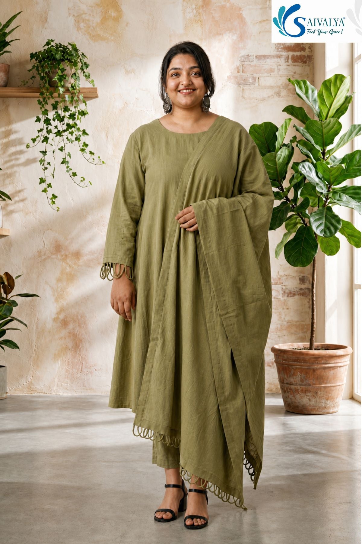 Front view of a smiling woman standing in a rustic room with potted plants, wearing the Olive Loop Grace Suit Set featuring an olive-green A-line kurta, straight pants, and a matching dupatta, all highlighted by distinct loop embroidery details along the sleeve cuffs and hems.