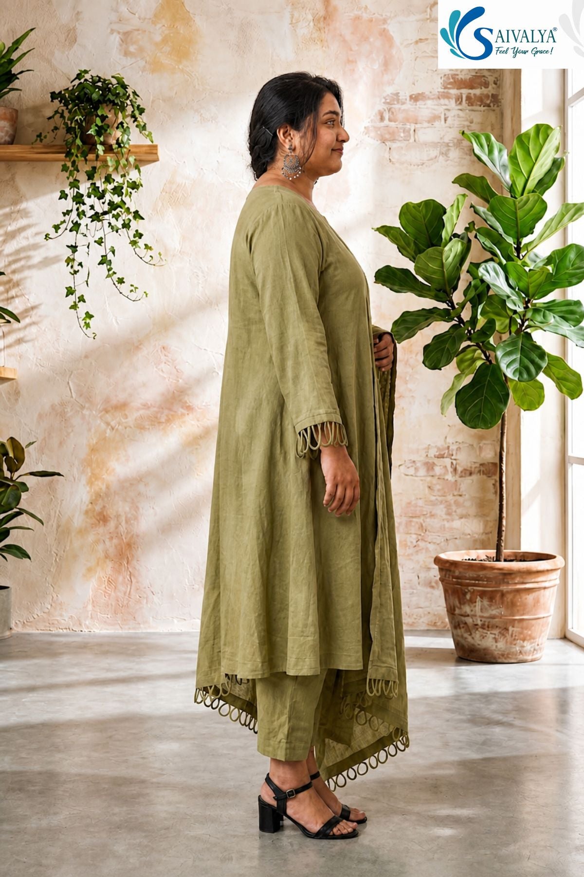 Side view of a woman standing in a rustic room with potted plants, wearing the Olive Loop Grace Suit Set showcasing the side profile of the flared olive-green A-line kurta, straight pants, and draped dupatta with distinct loop embroidery details.
