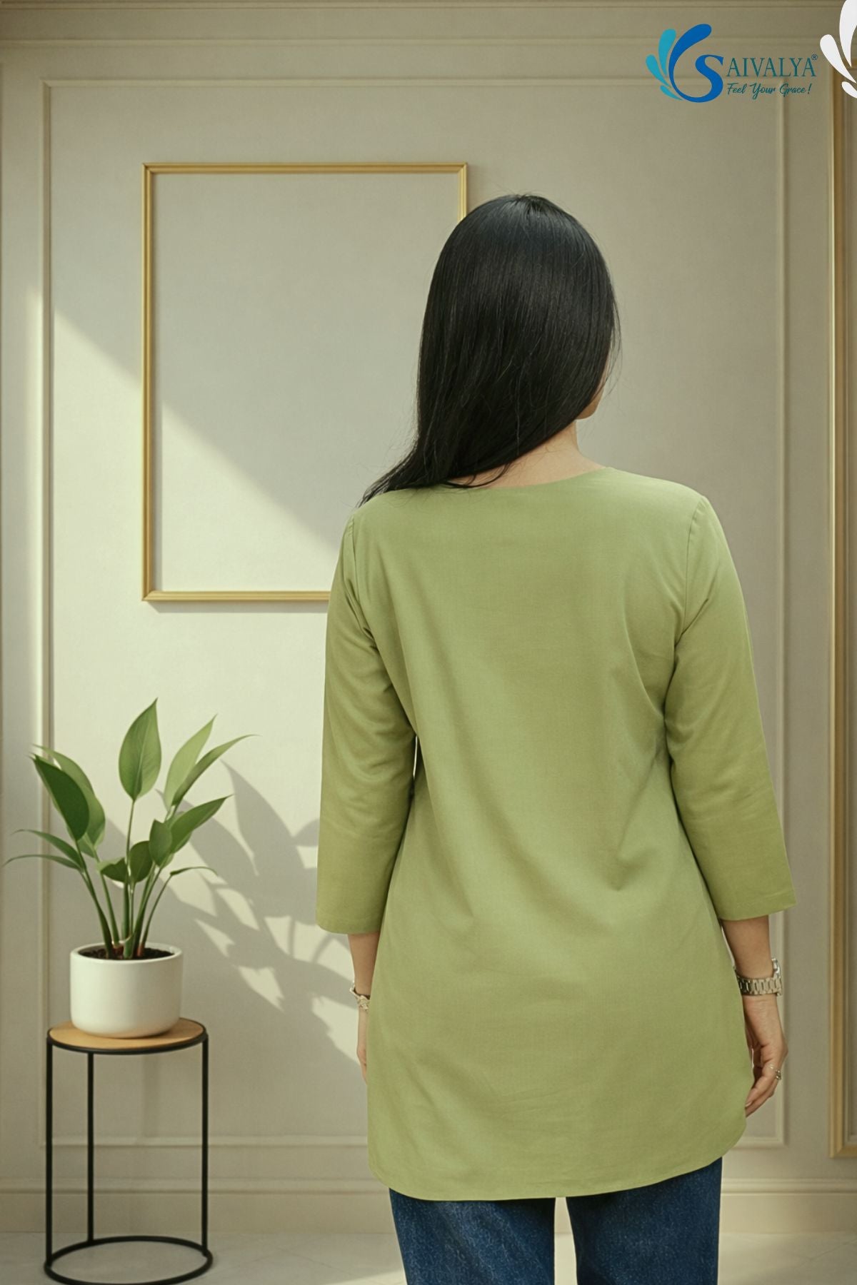 Full-length back view of a woman standing in a minimalist indoor studio setting with a gold frame and a potted plant in the background, wearing the Prakriti Sage Overlap Tunic highlighting its clean back profile, round neckline, and three-quarter sleeves, paired with blue denim jeans.