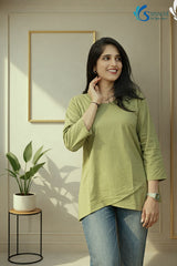 Three-quarter medium view of a smiling woman looking off-camera with a hand resting near her neck in a minimalist indoor studio setting with a gold frame and a potted plant in the background, wearing the Prakriti Sage Overlap Tunic highlighting its round neckline, three-quarter sleeves, and asymmetric overlap tulip hemline, paired with blue denim jeans.